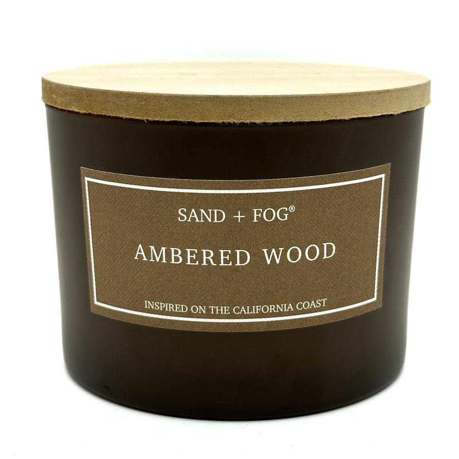 Candle with a label reading 'Sand + Fog Ambered Wood' on a white background