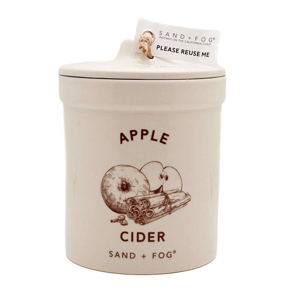 A ceramic jar candle by Sand + Fog in the Apple Cider scent, featuring a cream-colored body with a brown apple and cinnamon illustration and dark brown text, complete with a paper tag on the lid.