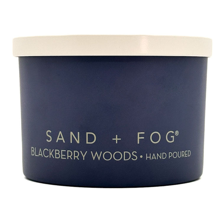 A dark navy blue cylindrical jar with a cream-coloured lid, labeled 'SAND + FOG BLACKBERRY WOODS HAND POURED' in light cream lettering, on a plain white background.
