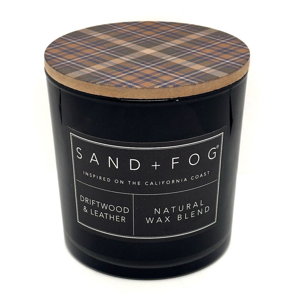 A black glass jar candle with a lid featuring a brown, beige, and blue plaid pattern. The front label reads 'SAND + FOG' and 'DRIFTWOOD & LEATHER NATURAL WAX BLEND'.