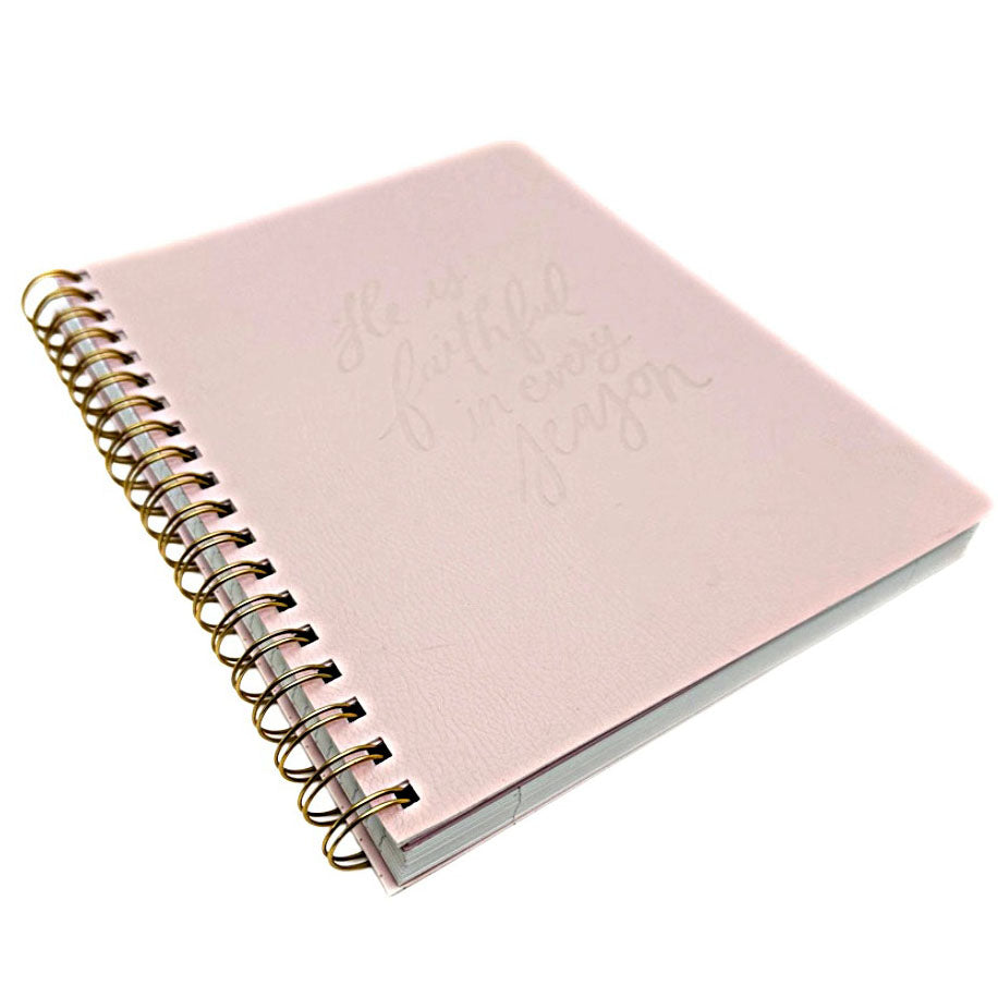 A light pink spiral-bound notebook with a gold wire coil binding and embossed cursive text on the cover 'He is faithful in every season' shown on a plain white background.