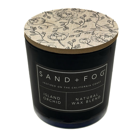 Island Orchid black candle with floral lid labeled 'Sand + Fog' on a white background