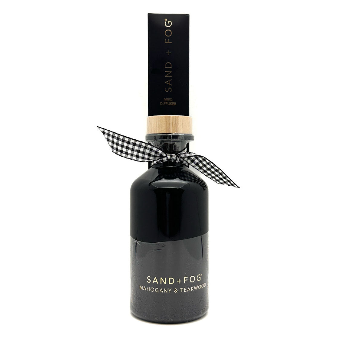 A dark glass bottle of Sand + Fog Mahogany & Teakwood fragrance with a black cap, a light wood collar, and a black-and-white gingham ribbon tied around the neck, on a white background.