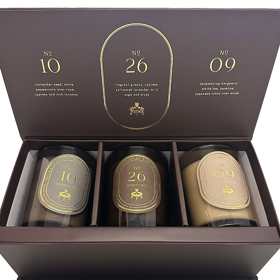 A luxury Ninety Six by DW Home three-candle gift set in a dark brown box with gold lettering. The open lid displays scents numbered 10, 26, and 09, and the box contains three dark glass candles with gold and beige labels.