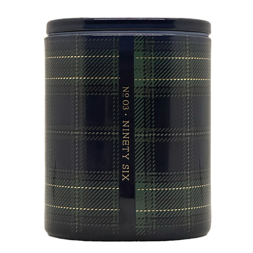 A cylindrical candle with a dark navy and green tartan plaid pattern, featuring a vertical black band with gold text reading 'Nº 03 · Ninety Six'.