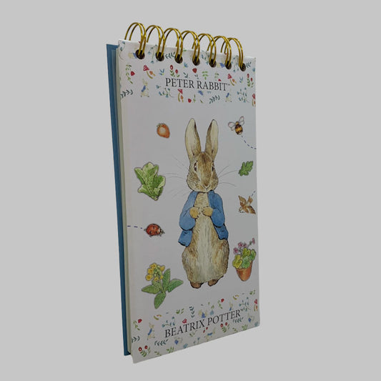 A Peter Rabbit spiral-bound notebook or desk pad with gold wire binding, featuring an illustration of Peter Rabbit in a blue jacket on a white background with floral details and the text 'PETER RABBIT' and 'BEATRIX POTTER'.