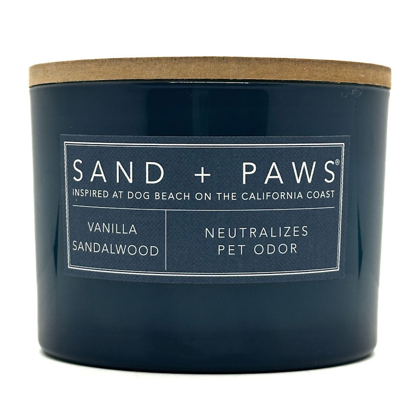 Vanilla Sandalwood Navy blue candle with a wooden lid labeled 'Sand + Paws' on a white background