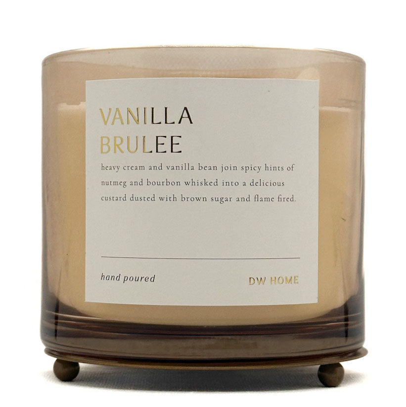 A candle in a clear glass jar with a beige wax fill and a white label reading 'VANILLA BRULEE' and 'DW HOME'. The jar sits on a dark brown wooden stand against a plain white background.