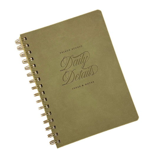 An olive green A5 spiral-bound notebook with gold-toned wire binding and embossed text reading 'PRINCE STUDIO Daily Details IDEAS & NOTES' on a white background.