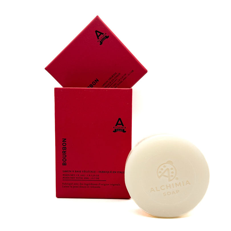 A round cream-colored bar of Alchimia Soap labeled 'Bourbon' is displayed next to its red cardboard packaging box and an open matching box on a clean white background.