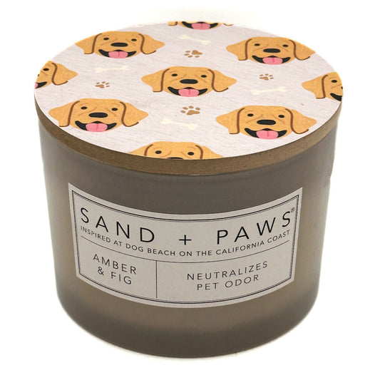 A cylindrical pet candle jar with a grey-brown body and a lid printed with a repeating pattern of yellow Labrador retriever faces and paw prints. The front label reads 'SAND + PAWS' and 'AMBER & FIG'.