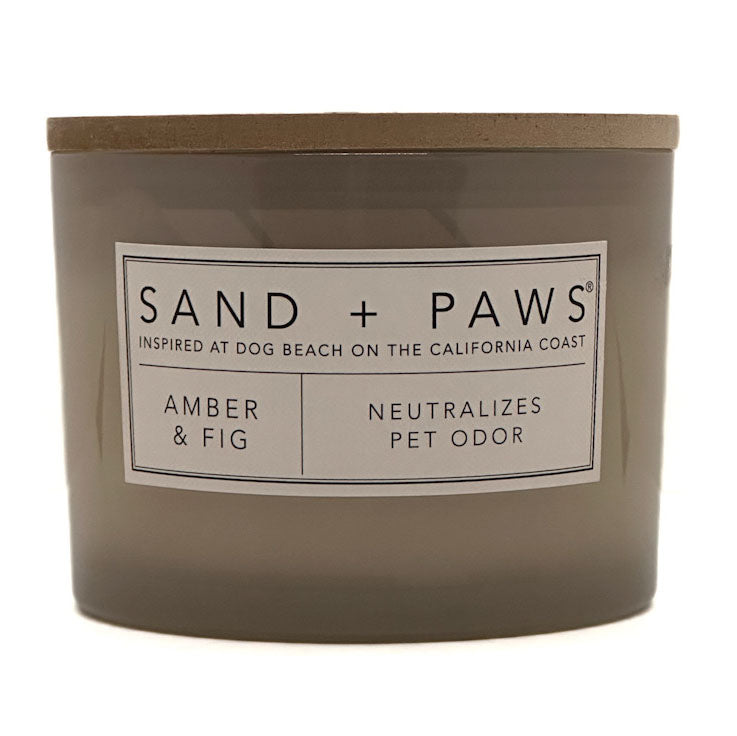 A jar candle labeled 'SAND + PAWS' in the scent 'Amber & Fig', with a taupe-brown glass container and lid on a white background.