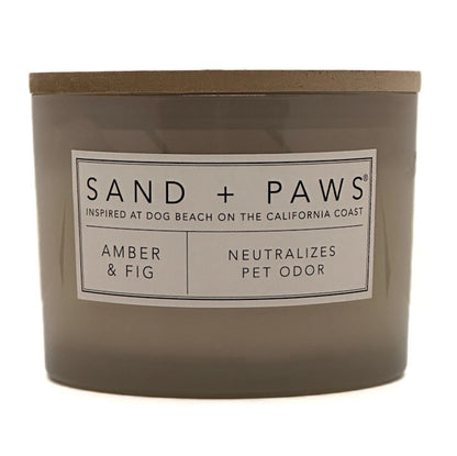 A jar candle labeled 'SAND + PAWS' in the scent 'Amber & Fig', with a taupe-brown glass container and lid on a white background.