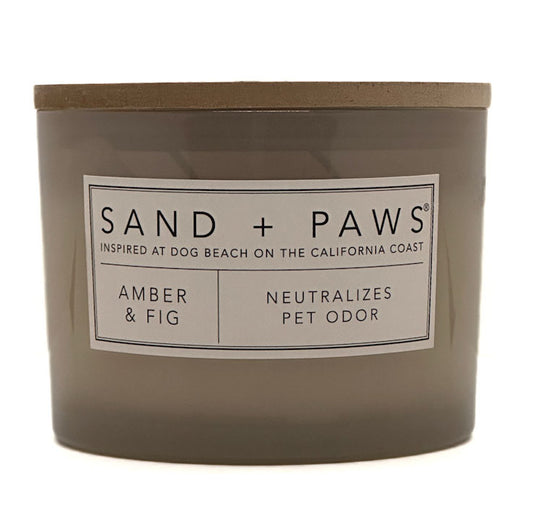 A jar candle labeled 'SAND + PAWS' in the scent 'Amber & Fig', with a taupe-brown glass container and lid on a white background.