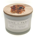 A jar candle with a light grey body and a round wooden lid featuring an illustrated golden retriever. The front label reads 'SAND + PAWS' and 'AMBER & FIG'.