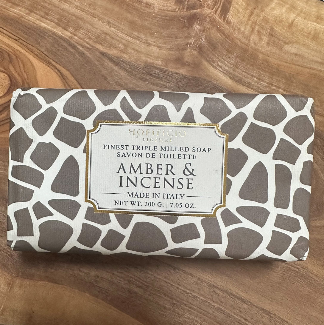 A rectangular bar of soap wrapped in a brown and white irregular patterned paper, with a central label reading 'Hoffmann Bros. Amber & Incense' and 'Finest Triple Milled Soap Made in Italy,' placed on a wooden surface.