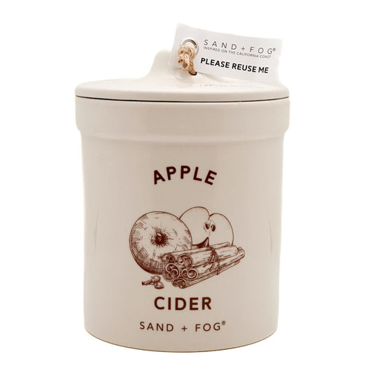 A ceramic jar candle by Sand + Fog in the Apple Cider scent, featuring a cream-colored body with a brown apple and cinnamon illustration and dark brown text, complete with a paper tag on the lid.