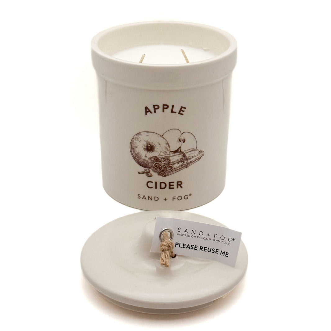 A white ceramic candle jar labeled 'APPLE CIDER' with a brown apple illustration, shown with its lid placed in front containing a small wick and a card that reads 'PLEASE REUSE ME.' The brand 'SAND + FOG' is visible on the label and tag.