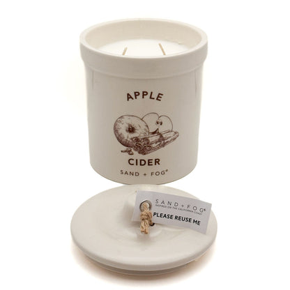 A white ceramic candle jar labeled 'APPLE CIDER' with a brown apple illustration, shown with its lid placed in front containing a small wick and a card that reads 'PLEASE REUSE ME.' The brand 'SAND + FOG' is visible on the label and tag.