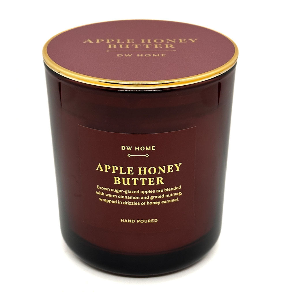 A DW Home Apple Honey Butter scented candle in a dark amber glass jar with a gold rim and a matching dark maroon lid, shown on a white background.