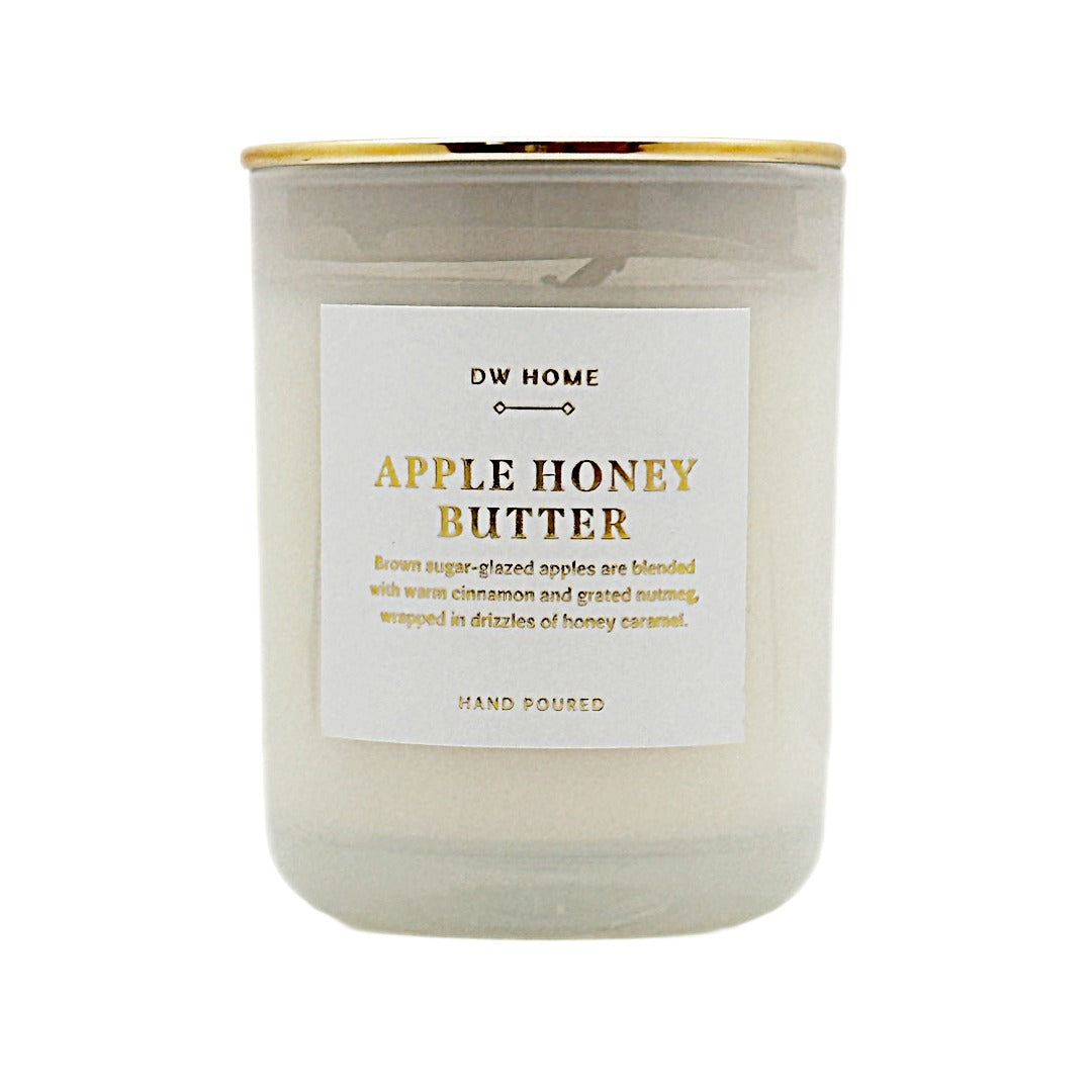 A glass jar candle with a white wax body, a white label reading 'DW HOME APPLE HONEY BUTTER,' and a gold metal lid, shown on a plain white background.