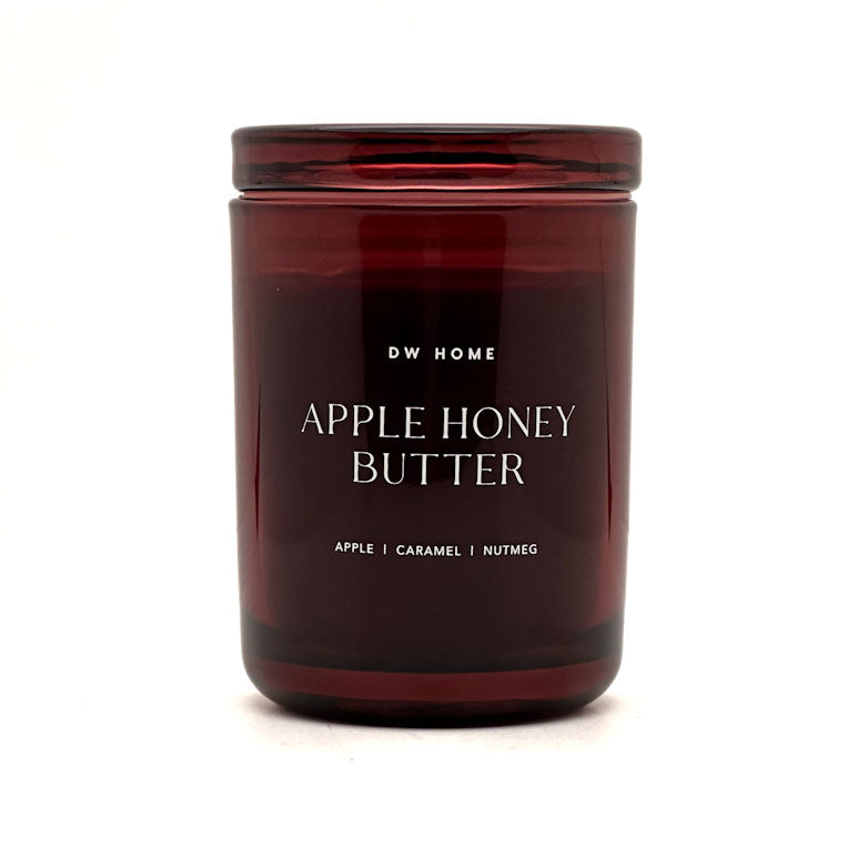 A dark amber glass jar candle with a matching lid, featuring white text that reads 'DW HOME APPLE HONEY BUTTER' and 'APPLE | CARAMEL | NUTMEG' on a clean white background.