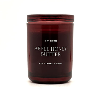 A dark amber glass jar candle with a matching lid, featuring white text that reads 'DW HOME APPLE HONEY BUTTER' and 'APPLE | CARAMEL | NUTMEG' on a clean white background.