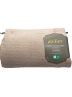 A folded beige chenille throw blanket with a ribbed texture, featuring a dark gray paper label that reads 'atelier luxury chenille throw oversized' on a white background.