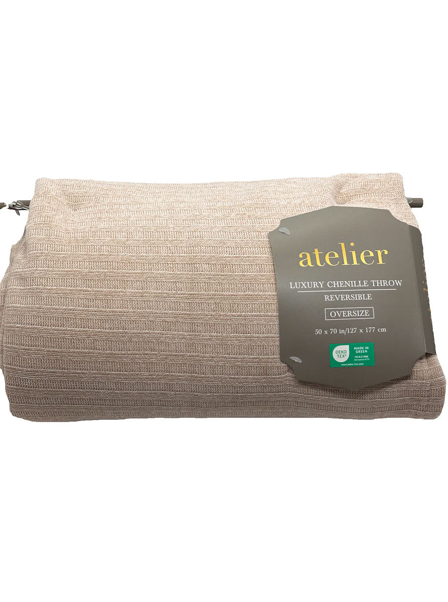 A folded beige chenille throw blanket with a ribbed texture, featuring a dark gray paper label that reads 'atelier luxury chenille throw oversized' on a white background.