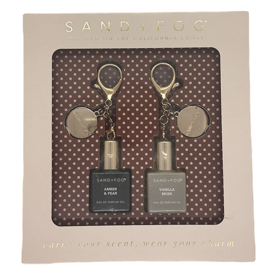 Sand + Fog keychain gift set in its original packaging, featuring two 10ml perfume oil vials (Amber & Pear and Vanilla Musk) with deer-head keychains on a brown polka-dot background insert.