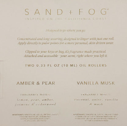A beige product card or packaging insert for Sand + Fog oil rollers, featuring scent names Amber & Pear and Vanilla Musk with descriptive text and scent notes in a gold-toned serif font.