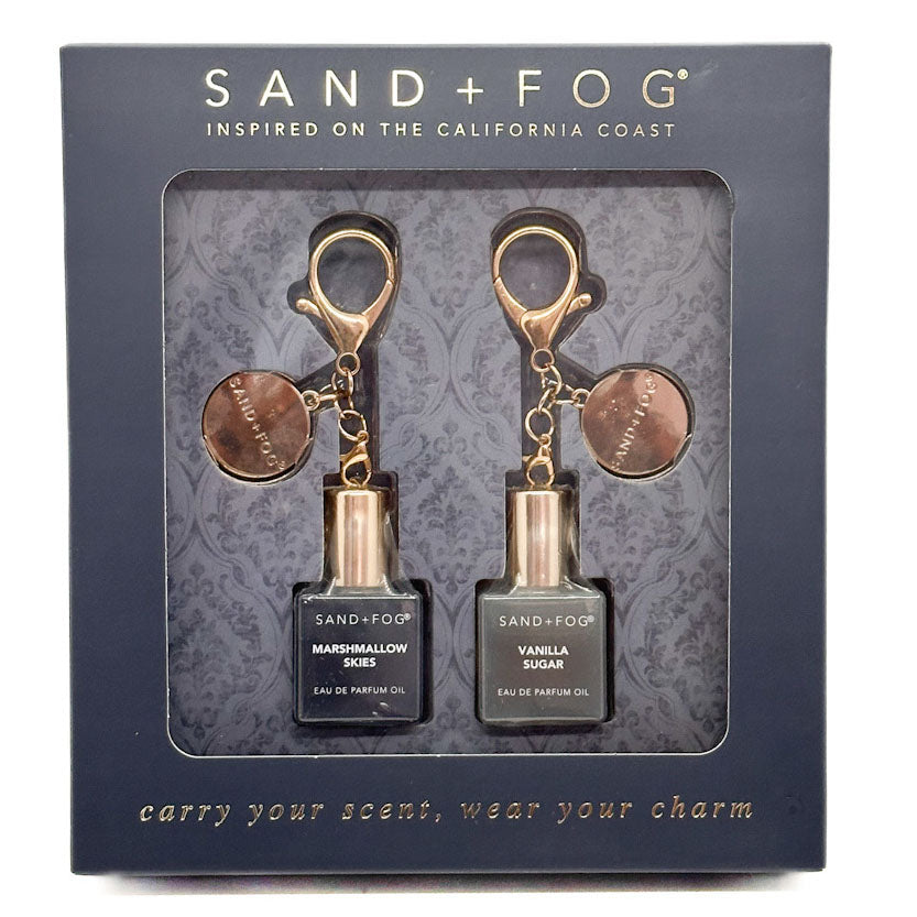 Sand + Fog perfume oil set in its original dark blue-gray packaging box, featuring two small perfume oil bottles with rose gold and bronze keychain-style caps labeled Marshmallow Ees and Vanilla Sugar.