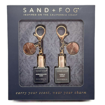 Sand + Fog perfume oil set in its original dark blue-gray packaging box, featuring two small perfume oil bottles with rose gold and bronze keychain-style caps labeled Marshmallow Ees and Vanilla Sugar.