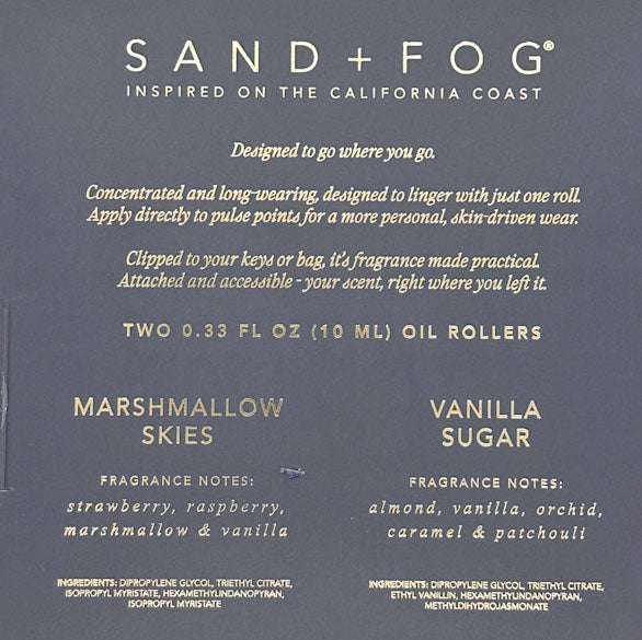 Back of a Sand + Fog oil roller package in a dark slate blue-gray color, showing product details, scent notes for Marshmallow Skies and Vanilla Sugar, and usage text in white and gold lettering.