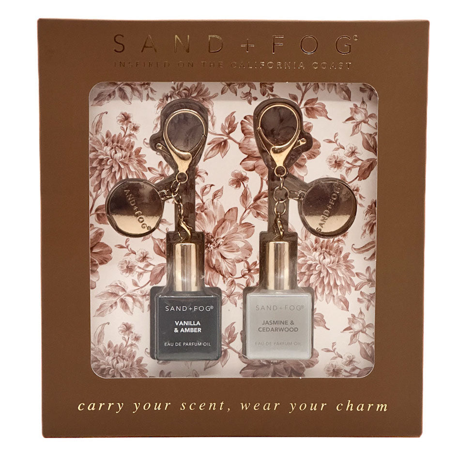 Sand + Fog carry your scent, wear your charm set in a brown floral gift box. The set includes a Vanilla & Amber vanilla perfume oil and a Jasmine & Cedarwood eau de parfum oil, each with a gold-toned keychain charm.