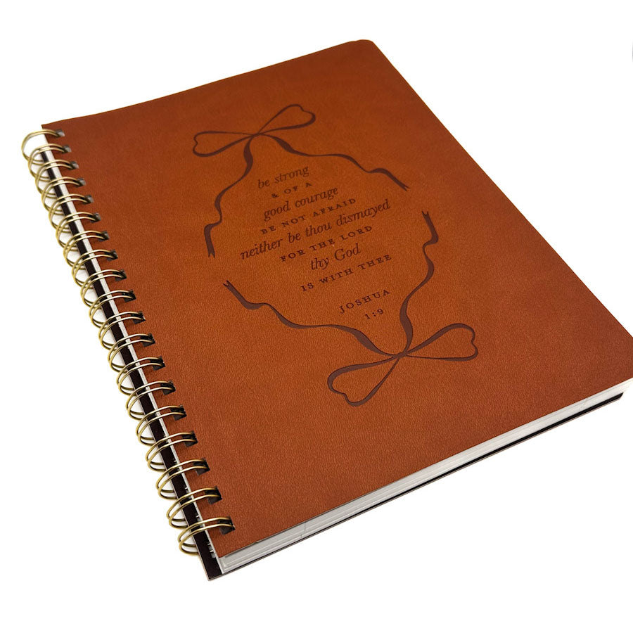 A brown leather-bound spiral notebook with a gold-tone wire binding and an embossed design on the cover featuring a bow and a Bible verse from Joshua 1:9.