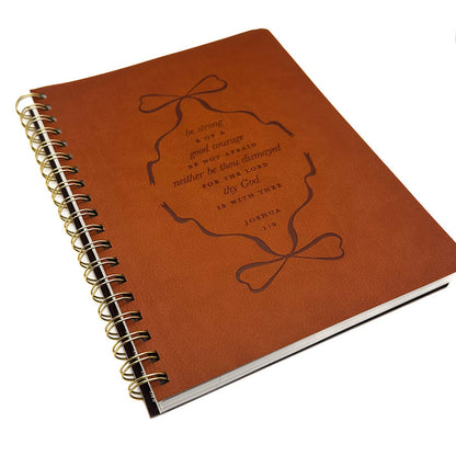 A brown leather-bound spiral notebook with a gold-tone wire binding and an embossed design on the cover featuring a bow and a Bible verse from Joshua 1:9.