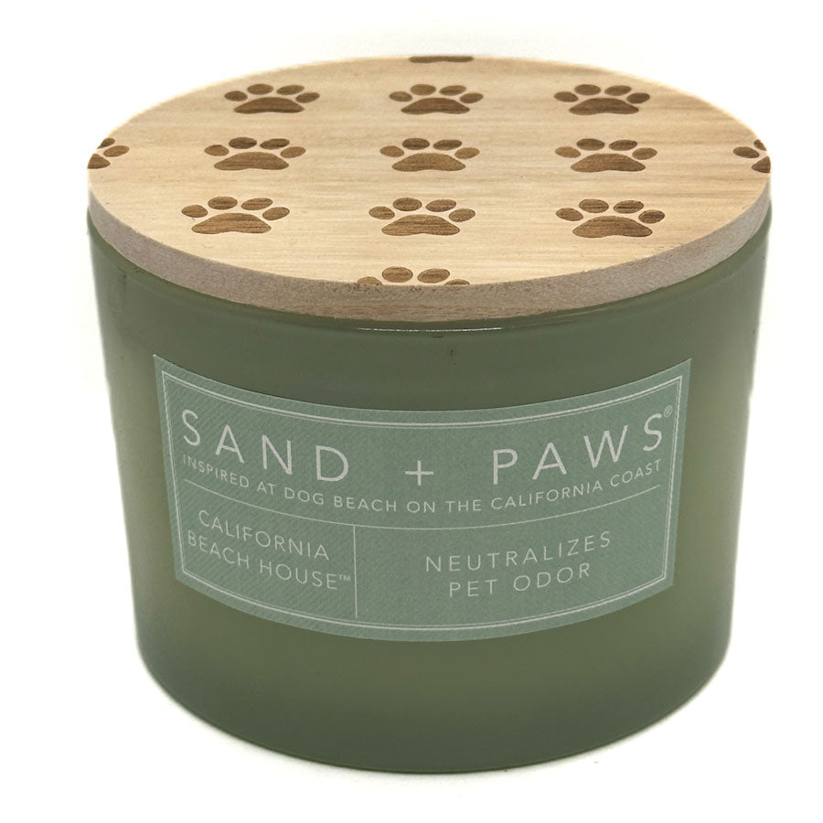 An olive green candle jar with a light wood lid featuring engraved paw prints, labeled 'SAND + PAWS' and 'California Beach House'.