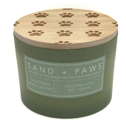 An olive green candle jar with a light wood lid featuring engraved paw prints, labeled 'SAND + PAWS' and 'California Beach House'.