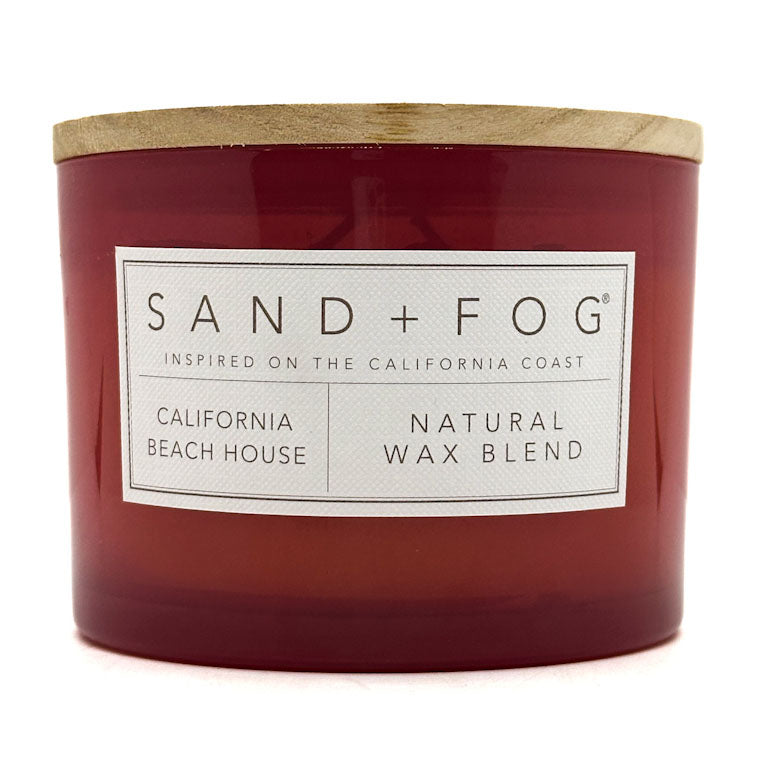 A red scented candle with a wooden lid on top. The label on the candle reads 'SAND + FOG California Beach House Natural Wax Blend'.