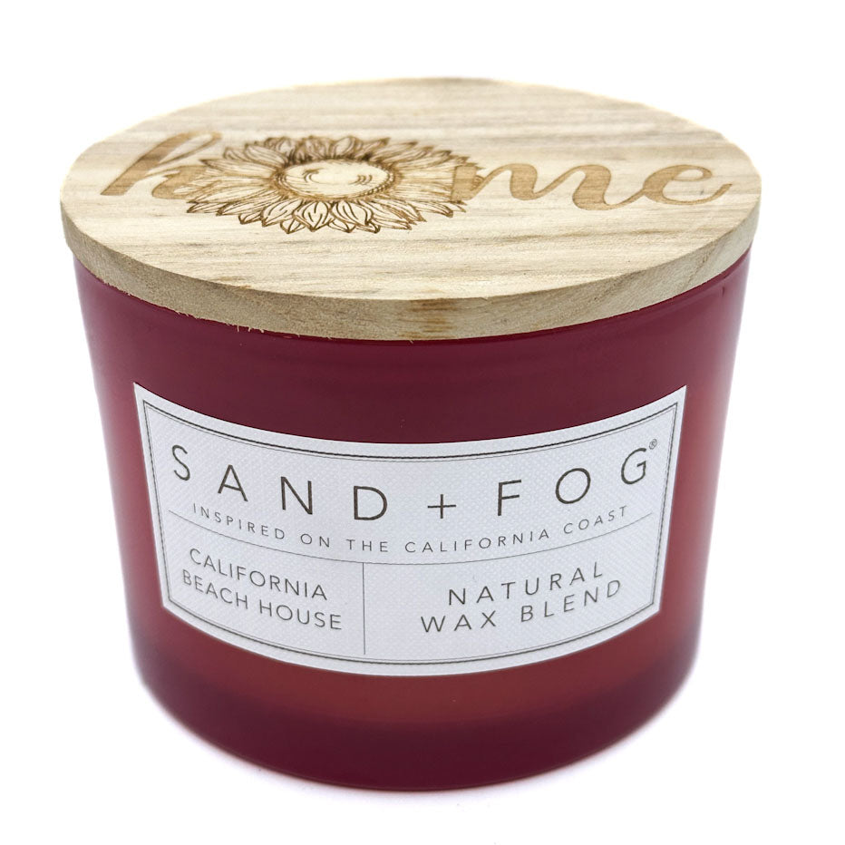 A red scented candle with a wooden lid on top, featuring an engraved 'home' and a sunflower. The label on the candle reads 'SAND + FOG California Beach House Natural Wax Blend'.