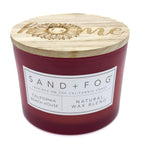 A red scented candle with a wooden lid on top, featuring an engraved 'home' and a sunflower. The label on the candle reads 'SAND + FOG California Beach House Natural Wax Blend'.