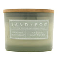 A jar candle with a translucent sage green glass body and a natural brown wood lid, featuring a white label that reads 'SAND + FOG' and 'CALIFORNIA BEACH HOUSE | NATURAL WAX BLEND'.