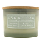 A jar candle with a translucent sage green glass body and a natural brown wood lid, featuring a white label that reads 'SAND + FOG' and 'CALIFORNIA BEACH HOUSE | NATURAL WAX BLEND'.
