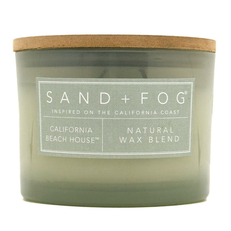 A jar candle with a translucent sage green glass body and a natural brown wood lid, featuring a white label that reads 'SAND + FOG' and 'CALIFORNIA BEACH HOUSE | NATURAL WAX BLEND'.
