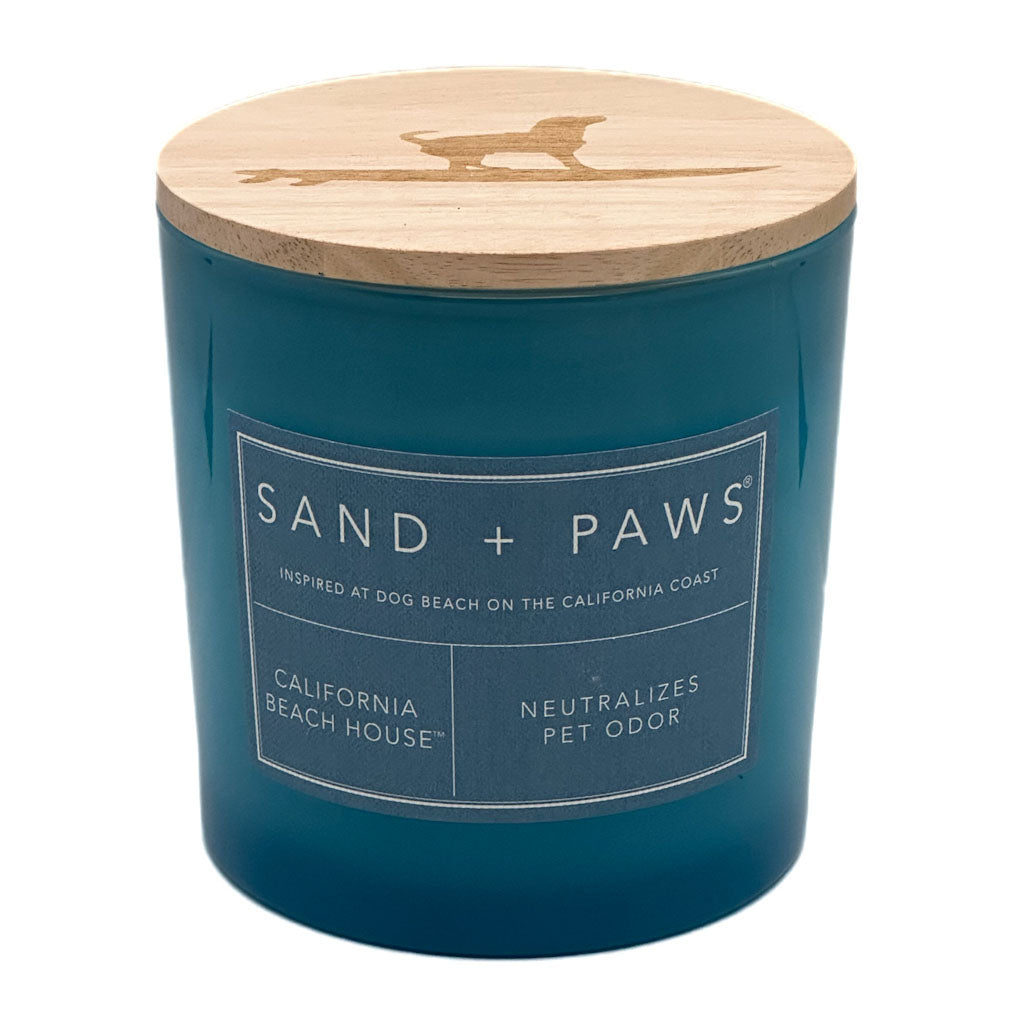 A teal blue jar candle with a light natural wood lid featuring a laser-cut dog silhouette. The front has a white label that reads 'SAND + PAWS' and 'CALIFORNIA BEACH HOUSE'.