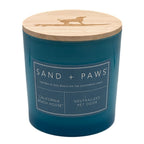 A teal blue jar candle with a light natural wood lid featuring a laser-cut dog silhouette. The front has a white label that reads 'SAND + PAWS' and 'CALIFORNIA BEACH HOUSE'.