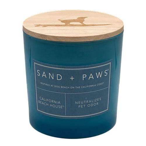 A teal blue jar candle with a light natural wood lid featuring a laser-cut dog silhouette. The front has a white label that reads 'SAND + PAWS' and 'CALIFORNIA BEACH HOUSE'.