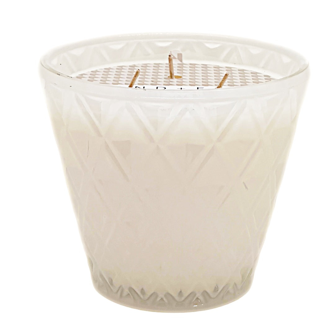 A California Beach House white wax candle in a faceted, diamond-patterned clear glass jar with three wicks, shown against a plain white background. SAND + FOG.