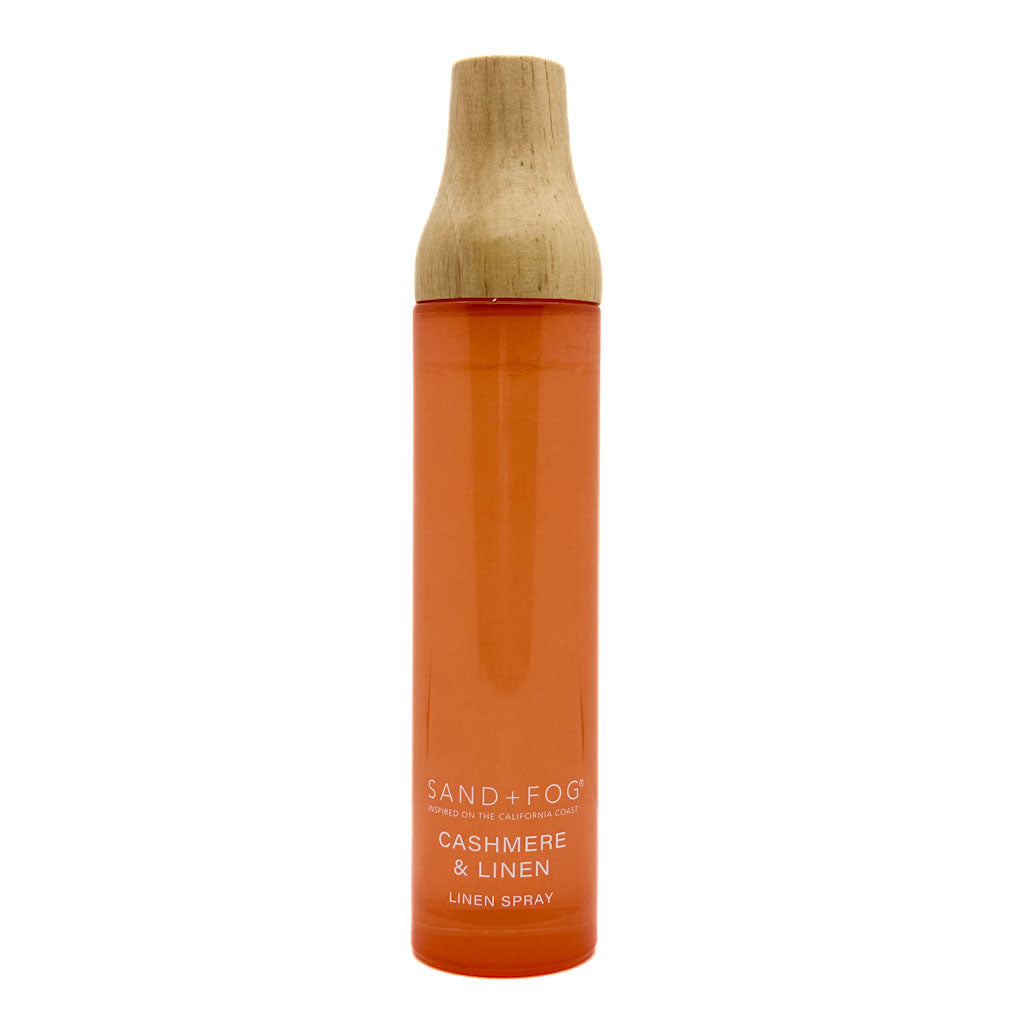 A tall cylindrical spray bottle of Sand + Fog Cashmere & Linen linen spray, featuring a translucent orange body and a natural light wood cap, set against a plain white background.